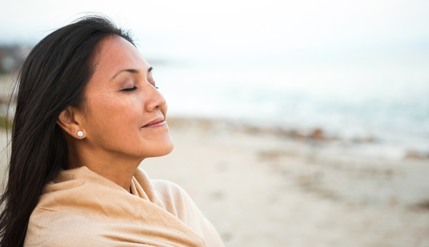 A woman wrapped in a shawl closes her eyes and smiles as she stands on the beach. 